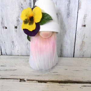 May include: A white ceramic pot gnome with a pink and white beard. The gnome is wearing a yellow pansy flower with purple accents.