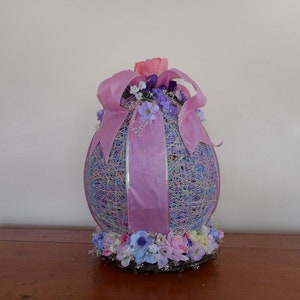 Puede incluir: Un huevo de Pascua grande, de color morado y azul pastel, decorado con una cinta rosa y un arreglo floral de flores rosas, moradas y blancas.
