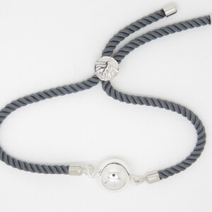 Peut inclure: Un bracelet en corde gris avec un charme argenté en forme ronde et texturée et un charme rond lisse plus petit. Le bracelet est réglable avec un nœud coulissant.