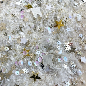 May include: Close-up of a collection of decorative elements, including gold, silver, and white stars, iridescent sequins, and white tooth-shaped cutouts. The items are scattered on a white, textured surface, creating a whimsical and festive appearance.