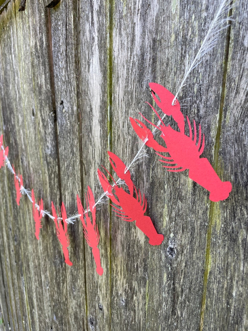 Peut inclure: Une guirlande de papier rouge en forme d'&eacute;crevisses avec une ficelle blanche suspendue &agrave; une cl&ocirc;ture en bois.