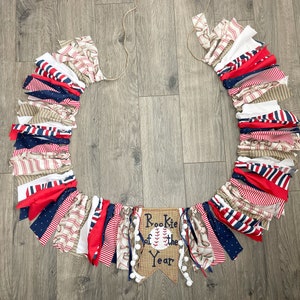 May include: A red, white, and blue fabric banner with a burlap banner that says "Rookie of the Year" and a baseball graphic. The fabric banner is made of strips of fabric with different patterns, including stripes, polka dots, and plaid.