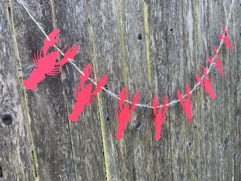 Peut inclure: Une guirlande de homards en papier rouge enfil&eacute;e sur une ficelle blanche, suspendue &agrave; une cl&ocirc;ture en bois vieilli.