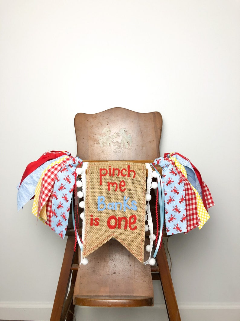 May include: A burlap banner with the text "Pinch me Banks is one" in red lettering. The banner is decorated with white pom-poms and is hanging from a high chair decorated with red, white, blue, and yellow ribbon.