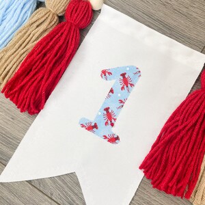 K&ouml;nnte beinhalten: Ein wei&szlig;es Stoffbanner mit einer blauen und roten Nummer eins mit einem Hummermuster. Das Banner ist mit roten, braunen und blauen Garnquasten und Holzperlen verziert.