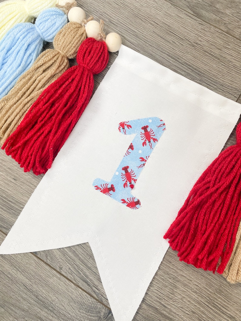 K&ouml;nnte beinhalten: Ein wei&szlig;es Stoffbanner mit einer blauen und roten Nummer eins mit einem Hummermuster. Das Banner ist mit roten, braunen und blauen Garnquasten und Holzperlen verziert.