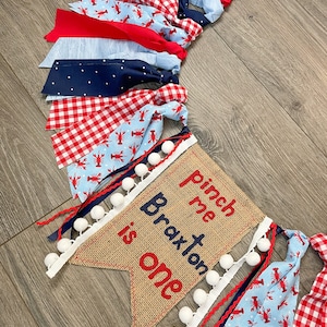 May include: A burlap banner with the text "Pinch me Braxton is one" in red and blue embroidery. The banner is decorated with white pom-poms and blue and red ribbon. The banner is part of a larger garland made of red and blue gingham fabric, blue fabric with white polka dots, and blue fabric with a lobster print.