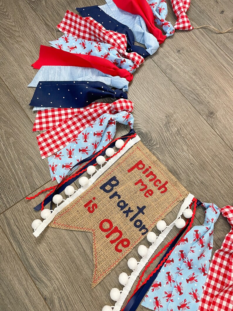 May include: A burlap banner with the text "Pinch me Braxton is one" in red and blue embroidery. The banner is decorated with white pom-poms and blue and red ribbon. The banner is part of a larger garland made of red and blue gingham fabric, blue fabric with white polka dots, and blue fabric with a lobster print.