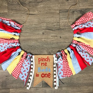 May include: A burlap banner with the text "pinch me Banks is one" in red and white lettering. The banner is decorated with white pom-poms and is surrounded by a garland of red, blue, yellow, and white fabric scraps with a lobster print.