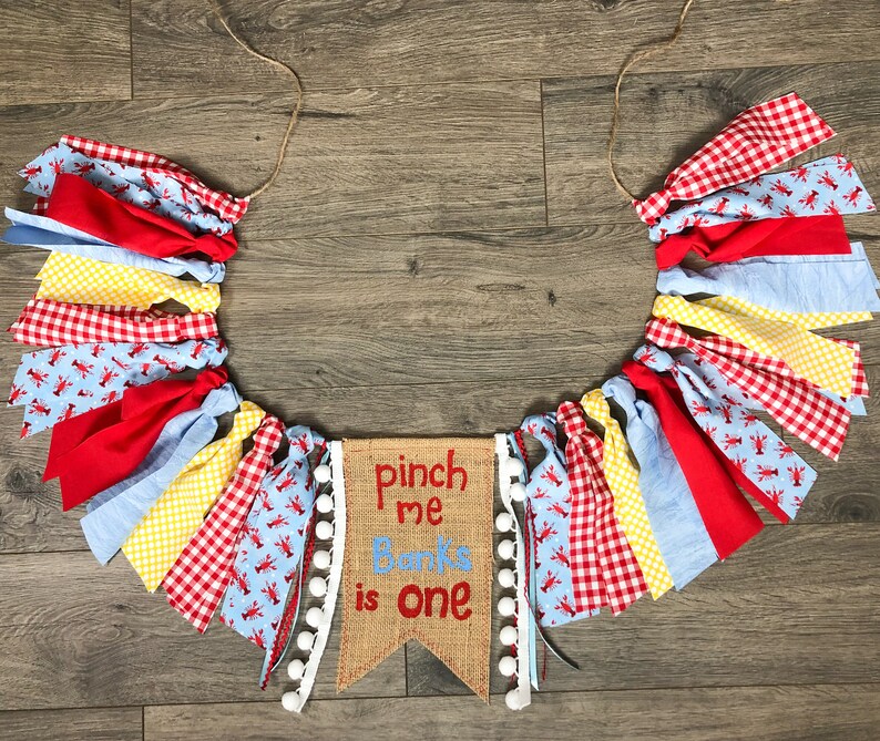May include: A burlap banner with the text "pinch me Banks is one" in red and white lettering. The banner is decorated with white pom-poms and is surrounded by a garland of red, blue, yellow, and white fabric scraps with a lobster print.