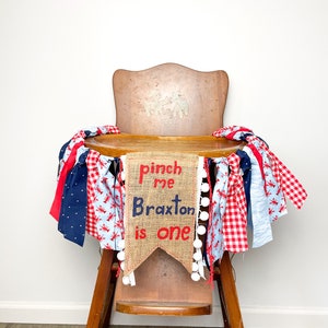 May include: A burlap banner with the text "Pinch me Braxton is one" in red and blue lettering, decorated with white pom-poms and red and blue gingham ribbon. The banner is draped over a wooden high chair.