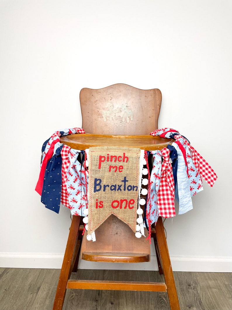May include: A burlap banner with the text "Pinch me Braxton is one" in red and blue lettering, decorated with white pom-poms and red and blue gingham ribbon. The banner is draped over a wooden high chair.