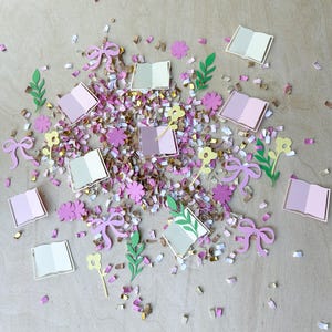 May include: A collection of paper cutouts including open books, bows, flowers, and leaves in shades of pink, green, and yellow. The cutouts are scattered among confetti in gold, pink, and white, creating a whimsical, decorative scene.