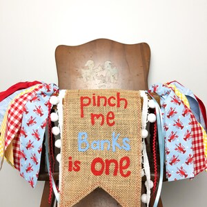 May include: A burlap banner with the text "Pinch me Banks is one" in red and blue lettering. The banner is decorated with white pom-poms and red and blue ribbon with a lobster print.