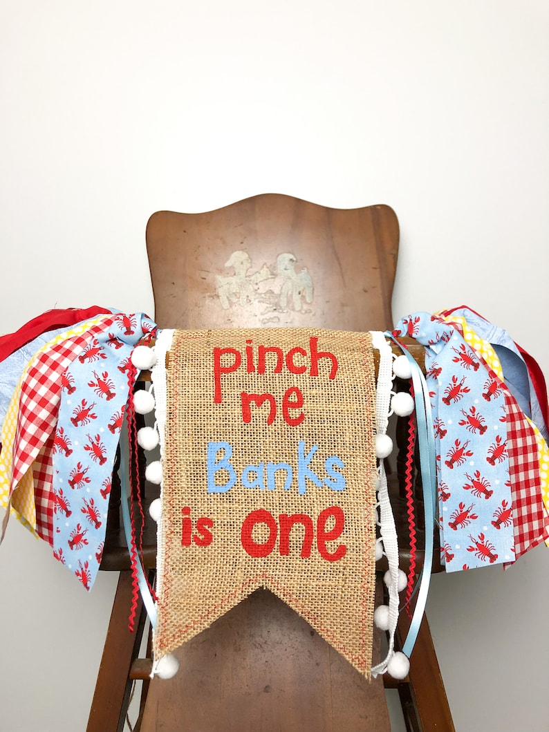 May include: A burlap banner with the text "Pinch me Banks is one" in red and blue lettering. The banner is decorated with white pom-poms and red and blue ribbon with a lobster print.