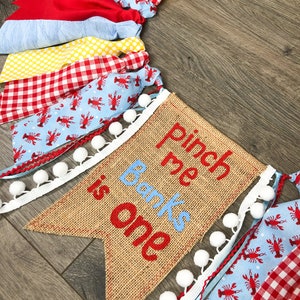 May include: A burlap banner with the text "Pinch me Banks is one" in red and blue embroidery. The banner is decorated with white pom-poms and a blue and white ribbon. The banner is surrounded by a garland of red, white, and blue fabric scraps.