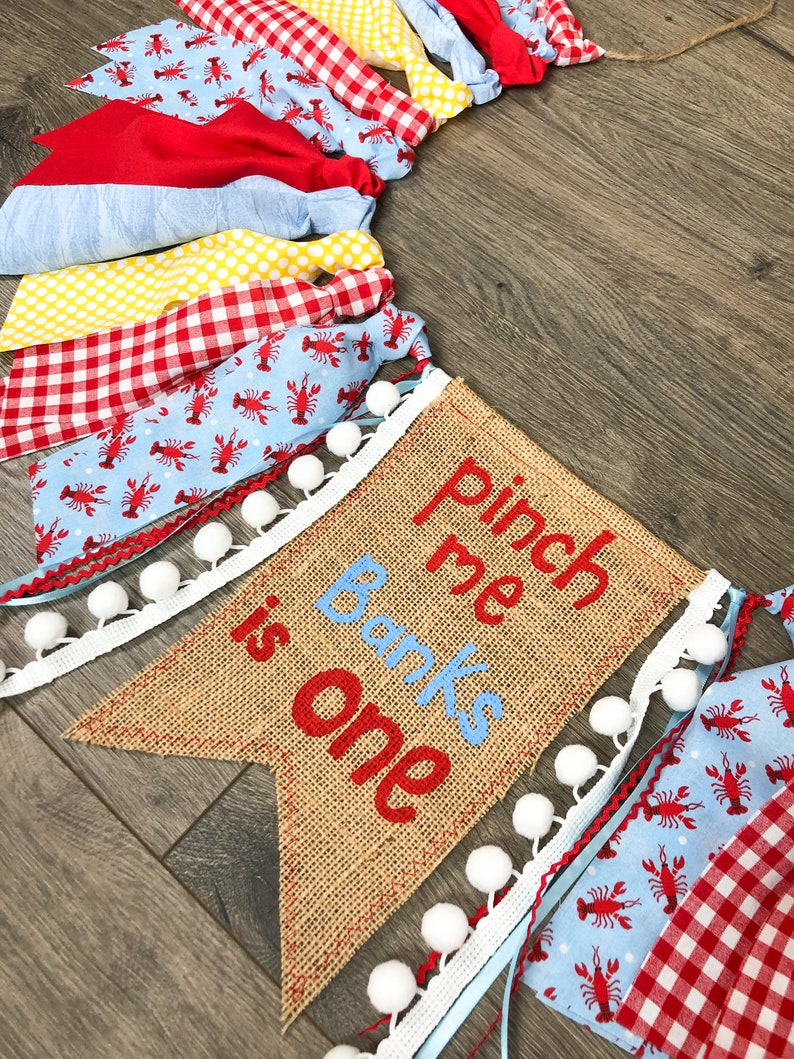 May include: A burlap banner with the text "Pinch me Banks is one" in red and blue embroidery. The banner is decorated with white pom-poms and a blue and white ribbon. The banner is surrounded by a garland of red, white, and blue fabric scraps.
