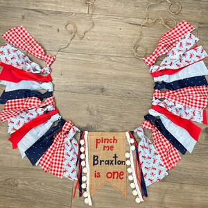 May include: A red, white, and blue fabric banner with lobster designs and white pom-poms. The banner says "Pinch me Braxton is one" on a burlap background.