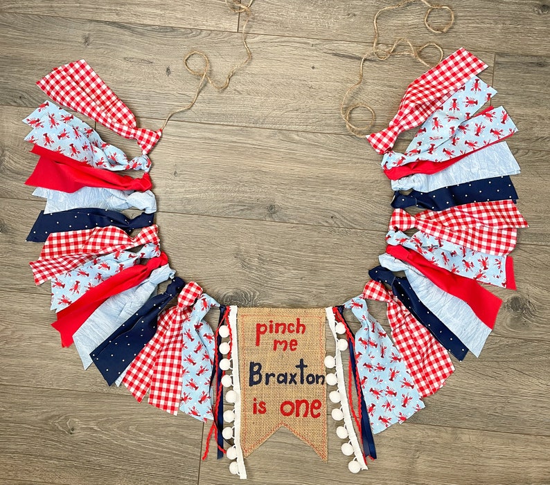 May include: A red, white, and blue fabric banner with lobster designs and white pom-poms. The banner says "Pinch me Braxton is one" on a burlap background.