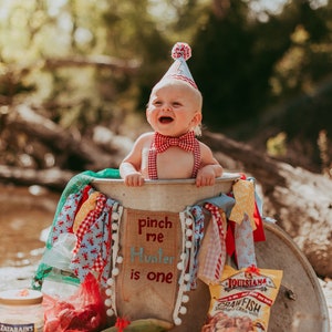May include: A baby boy wearing a red and white bow tie and a red and white party hat sits in a metal tub decorated with red, white, and blue fabric. The tub has a sign that says "pinch me Hunter is one".