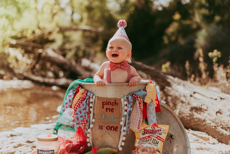 May include: A baby boy wearing a red and white bow tie and a red and white party hat sits in a metal tub decorated with red, white, and blue fabric. The tub has a sign that says "pinch me Hunter is one".