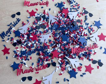 May include: A collection of red, white, and blue paper confetti featuring die-cut shapes of fighter jets, stars, sunglasses, and the word Maverick in script font, scattered across a light-colored surface.