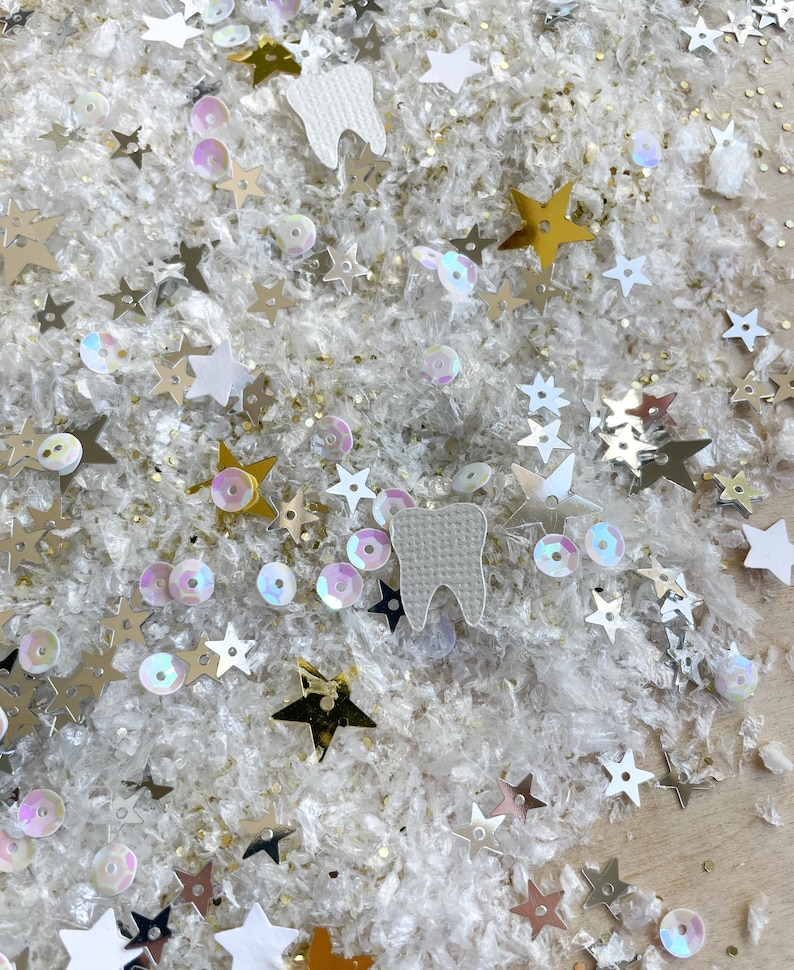 May include: Close-up of a collection of decorative elements, including gold, silver, and white stars, iridescent sequins, and white tooth-shaped cutouts. The items are scattered on a white, textured surface, creating a whimsical and festive appearance.