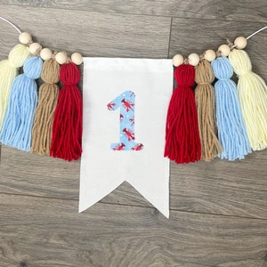 K&ouml;nnte beinhalten: Ein wei&szlig;es Banner mit der Zahl "1" in Rot und Blau mit einem Hummermuster. Das Banner ist mit einer Schnur aus Holzperlen und bunten Garnquasten in Gelb, Blau, Braun und Rot verziert.