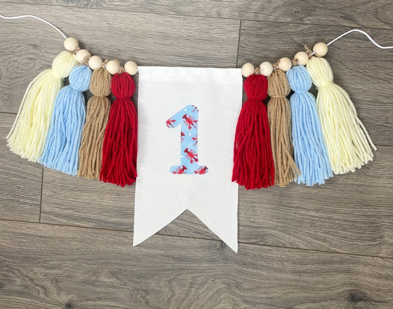 K&ouml;nnte beinhalten: Ein wei&szlig;es Banner mit der Zahl "1" in Rot und Blau mit einem Hummermuster. Das Banner ist mit einer Schnur aus Holzperlen und bunten Garnquasten in Gelb, Blau, Braun und Rot verziert.