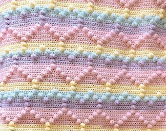 May include: A colorful crochet baby blanket with a chevron pattern and textured bobbles. The blanket is made in shades of pink, yellow, and blue.