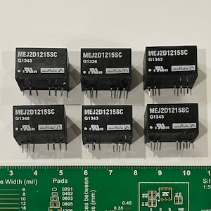 Może przedstawiać: Sześć czarnych komponentów elektronicznych oznaczonych "MEJ2D1215SC" z różnymi numerami G, "muRata Ps" i "cus" nadrukowanymi na nich. Komponenty są ułożone w dwóch rzędach po trzy, a leżą na zielonej linijce z oznaczeniami w calach i milimetrach.
