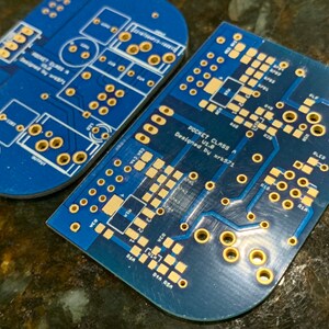 May include: Two blue circuit boards with gold-colored solder points. The top board has the text "POCKET CLASS A" and "Designed by xrk571".