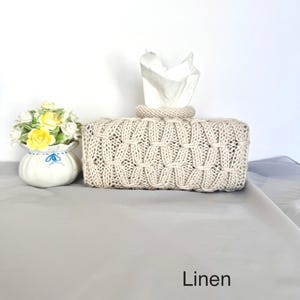 May include: A beige knitted tissue box cover with a textured pattern. White tissues are pulled from the top. A small white vase with yellow and white flowers is to the left. The word "Linen" is at the bottom.