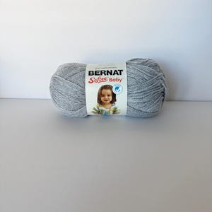 May include: A skein of Bernat Softee Baby yarn in a light gray color. The yarn is wrapped with a label featuring the brand name, product details, and a picture of a baby. The yarn is designed for baby projects.