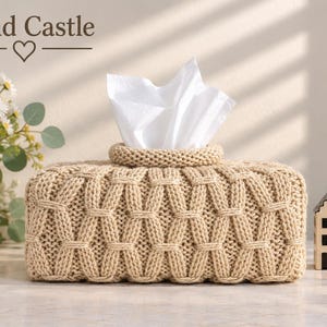 May include: A rectangular beige knit tissue box cover with a textured cable pattern, holding white tissues. To the left is a white vase with daisies, and to the right are two small house-shaped figurines. The text Sand Castle with a heart is visible.