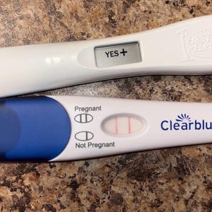 Puede incluir: Dos pruebas de embarazo de plástico blanco. La prueba superior muestra el texto YES+ en una pantalla digital. La prueba inferior muestra dos líneas rosas verticales junto a la palabra Pregnant y la marca Clearblue en azul.