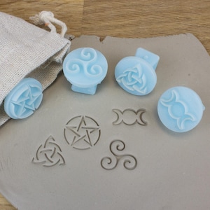 May include: A selection of light blue soap stamps and clay stamps featuring Wiccan and Celtic symbols. The stamps are displayed on a grey surface, with a linen bag visible. Symbols include a pentacle, triple moon, and triskelion.