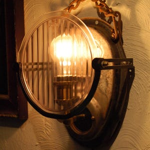 May include: A vintage-style wall lamp made from a repurposed Ford headlight. The lamp features a clear, ribbed glass lens, a visible lightbulb, and a metal frame with a chain for hanging. The lamp is mounted on a textured wall.