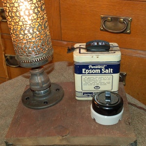 May include: A rustic lamp made from a pipe fitting and a decorative metal shade. The lamp is sitting on a wooden base with a vintage tin of Epsom salt and a black and white light switch.