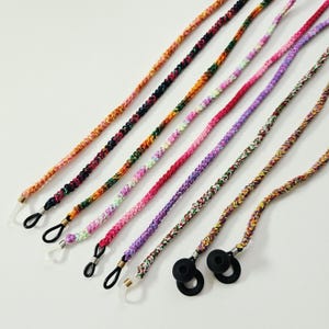 May include: A collection of eight braided cords for eyewear, featuring various multicoloured patterns including orange, pink, purple, green, and yellow. Each cord is finished with small metal coils and flexible loops for attaching to glasses frames.