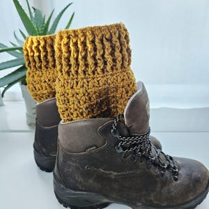 Puede incluir: Botas de senderismo marrones con polainas de ganchillo amarillo mostaza. Las botas tienen cordones blancos y negros. Están sobre un fondo blanco con una planta verde.