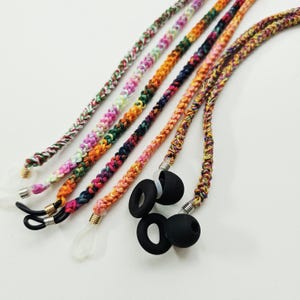 May include: A collection of braided eyeglass chains featuring multicolored yarn in various patterns, including pink, green, orange, and blue. Each cord is finished with metallic coils and flexible silicone loops in black or clear for securing eyewear.