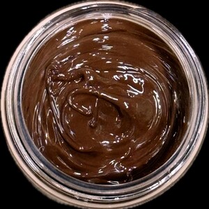 May include: A jar filled with a smooth, dark brown substance, possibly a cosmetic product or food item. The contents have a glossy appearance, reflecting light. The jar is clear, with a silver rim, set against a black background.