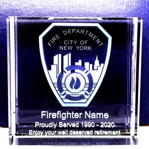 May include: A clear glass paperweight with an engraved image of the New York City Fire Department logo. The text "Firefighter Name Proudly Served 1990 - 2020 Enjoy your well deserved retirement" is engraved below the logo.