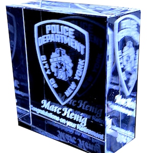 May include: A crystal paperweight with a 3D engraved image of the New York City Police Department logo. The text "Marc Henig" and "Congratulations on your Retirement" are also engraved on the paperweight.