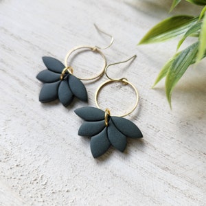 May include: A pair of dark gray, leaf-shaped earrings with gold-colored hoop accents. The earrings are displayed on a white, textured surface with a green plant in the background. The earrings have a modern, minimalist design.
