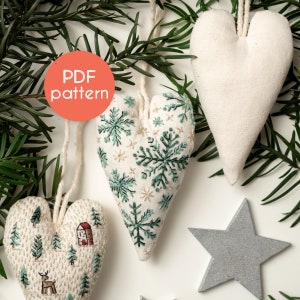 Puede incluir: Tres adornos de corazón de fieltro blanco con diferentes diseños de bordado. El primer corazón tiene un diseño de copo de nieve verde, el segundo corazón tiene una escena de bosque verde con una pequeña casa, y el tercer corazón es liso. Los adornos están colgados de un cordel blanco y están rodeados de ramas de pino verdes. El texto "PDF pattern" está en un círculo rojo.