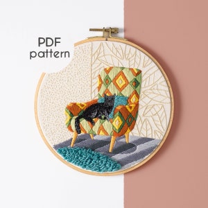 May include: Embroidered hoop art featuring a black cat lounging in a colorful, geometric patterned armchair. The background is a cream-colored fabric with a subtle leaf pattern.