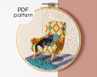 Op de afbeelding: Borduurwerk in een hoepel met een zwarte kat die in een kleurrijke, geometrisch gepatroneerde fauteuil ligt. De achtergrond is een crèmekleurige stof met een subtiel bladmotief.