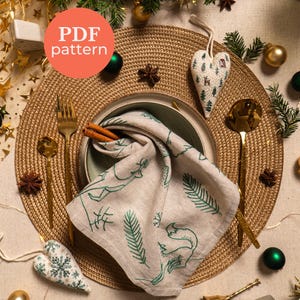 Puede incluir: Una mesa festiva con un mantel individual tejido, cubiertos dorados y una servilleta de lino con diseños bordados en verde. Adornos en forma de corazón y ramitas de hoja perenne se suman al tema navideño. La imagen incluye el texto "PDF pattern".